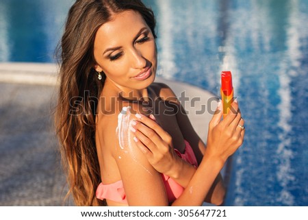 Sexy smiling woman with tanned skin is wearing bikini resting near swimming pool with sunscreen lotion, skin care