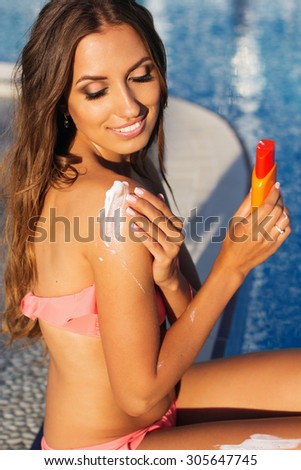Sexy smiling woman with tanned skin is wearing bikini resting near swimming pool with sunscreen lotion, skin care