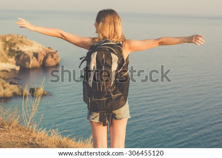 Back view traveler girl is standing on rock edge with backpack over sea view with backpack, summer time