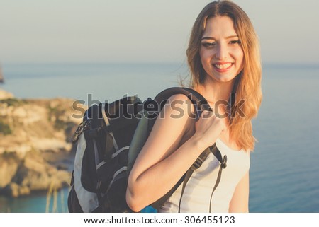 Pretty happy traveler girl is standing on rock edge with backpack over sea view with backpack, summer time