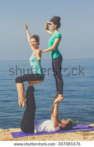 Young sportive trio group boy and two girls are practicing acroyoga exercises in the early morning on the beach over sea background