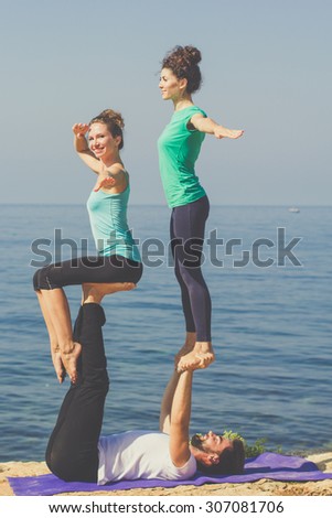 Young sportive trio group boy and two girls are practicing acroyoga exercises in the early morning on the beach over sea background
