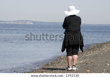 An older woman is looking out to sea at the beach with a big white hat on her head.