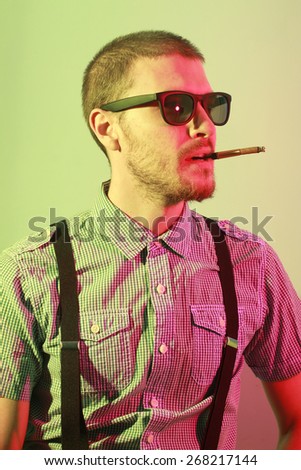 Portrait of handsome casual stylish young man with sunglasses, plaid short sleeve shirt and braces smoking cigarette with holder in green and red colored lights