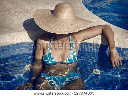Lady with straw hat and blue bikini in the pool, taking a suntan and enjoying the cold water of pool in a summer vacation.