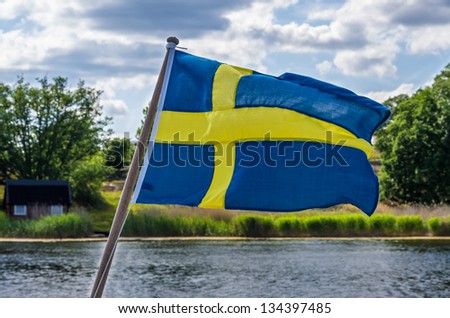 Swedish flag on a boat with a coastal landscape in background.