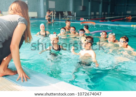Female instructor teaches children how to swim. Kids with goggles in water listening trainer. Happy kids in modern sport center. Concept of fun, leisure and recreation.