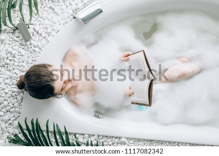 Woman lying in bath with foam and reads magazine