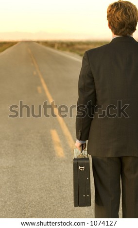 Business man standing in the middle of the road, facing towards the end of the road, in a black suit, with a briefcase, in the middle of the desert.