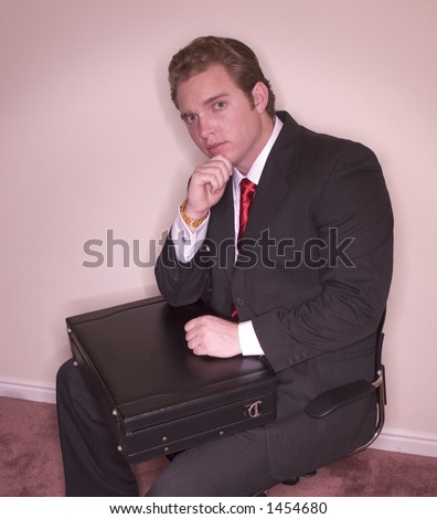 Business man is dressed in black suit, white shirt, and red tie, holding his chin and resting his hand on his briefcase with a sophisticated look