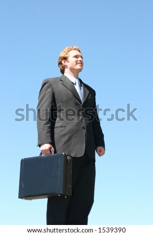 Business man in black suit in a forward looking pose carrying a briefcase against a blue sky