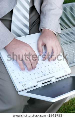Business man in tan shirt is typing on his white laptop as he is sitting on a park bench