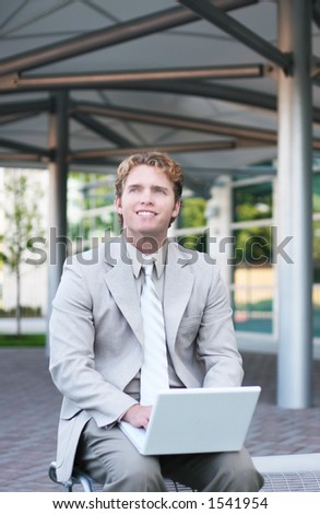 Business man in tan suit is sitting on park bench typing his white laptop and smiling