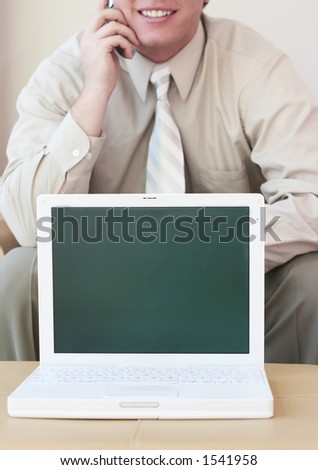 Business man wearing tan shirt is talking on his cell phone as laptop is displayed in front of him against tan background