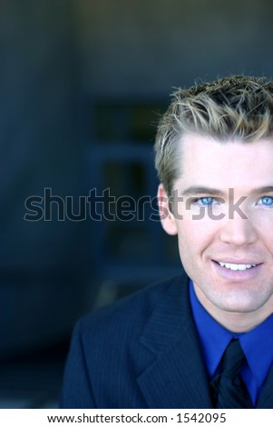 Blond hair, blue eye business man is wearing a dark blue suit with blue shirt and black tie is looking directly at you
