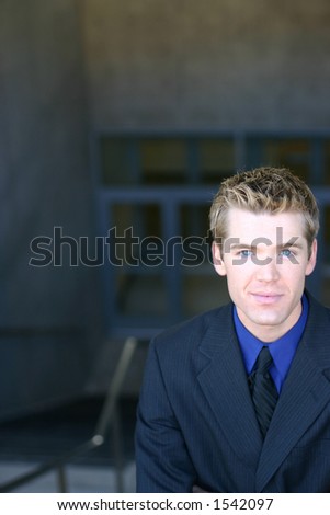 Blond hair blue eye business man is wearing a dark blue suit, black tie, and blue shirt is looking straight at you at the top of the stairs
