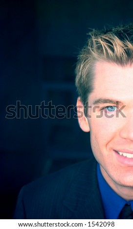 Blond hair, blue eye business man is wearing a blue suit with a blue shirt, and a black tie, as he is looking directly at you