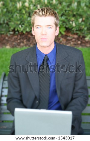 Blond hair blue  eye busines man is typing on his laptop on a park bench next to the green grass
