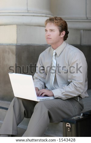 Business man in tan shirt and tie is looking up as his hands are on his white laptop, as he sits on the steps of the courthouse next to his briefcase