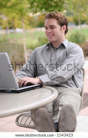 Business man wearing jacket types on his laptop with his feet up on a chair with trees and grass in the background