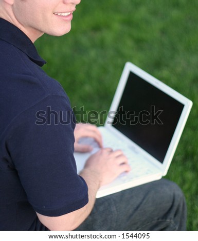 man dressed in blue shirt and jeans types on white laptop in the green grass