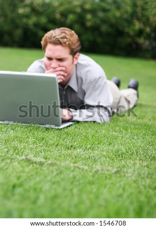 Businessman in gray shirt places hand over mouth as he thinks about what is on his laptop as he relaxes in the green grass