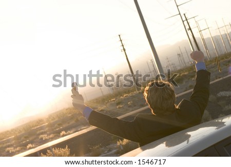 Businessman in black suit holds up cell phone in hand as he raises both his arms in victory as he stands outside his parked car in the middle of the desert as the sun is going down