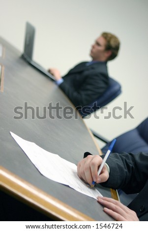 Businessman types on his laptop while another person signs a contract on a conference table