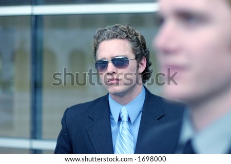 Business man in blue shirt and blue tie is wearing sunglasses with blurred shot of another business man in foreground