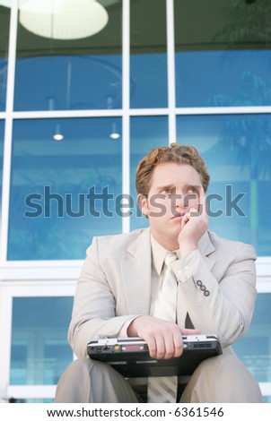 One young businessman sitting down holding laptop on his knees in front of office building with one hand on chin