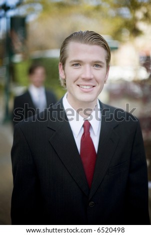portrait of one business man dressed in full suit with red tie with another businessman in full suit in background