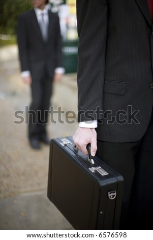 close up of businessman\'s hand holding briefcase with another businessman standing in background