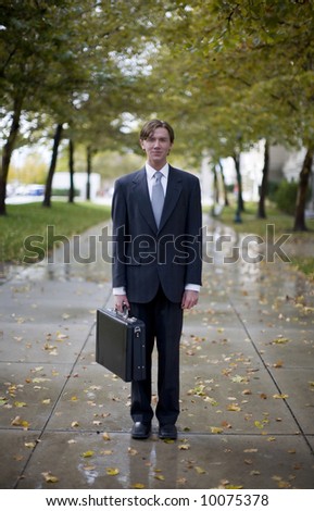 iconic single businessman wearing suit and tie holding briefcase standing in middle of walkway that is lined with trees