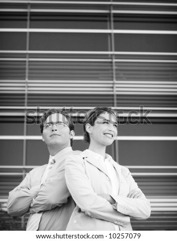 front view of businessman and businesswoman standing back to back with arms crossed in front of blue office building