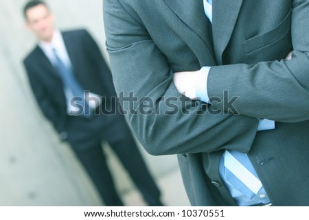 two businessmen one standing in front of the other with the first folding his arms and the second with hands in pockets