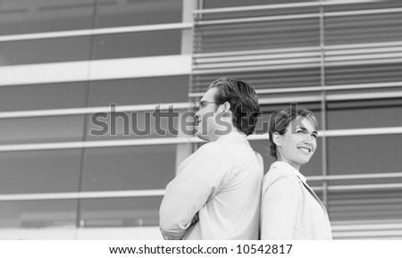 front view of businessman and businesswoman standing back to back with arms crossed in front of blue office building