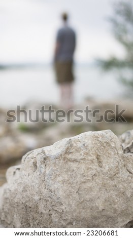 a distant man stands alone on the shore next to a large lake with a big rock as the focus in the foreground