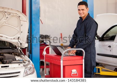 Happy young mechanic using a laptop computer and working on a car at an auto shop
