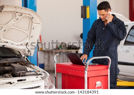 Handsome young mechanic talking to a customer on a cellphone while working on a computer at an auto shop