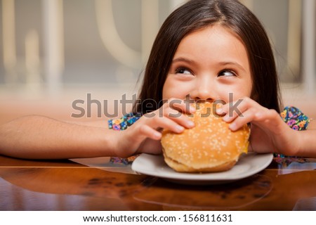 Cute Little Latin Girl Eating A Hamburger And Looking Up Towards Copy Space