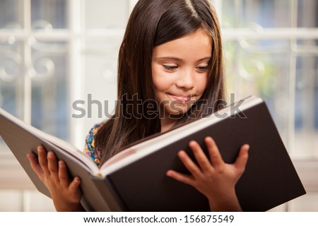 Cute little Hispanic girl reading a story from a big book in the living room