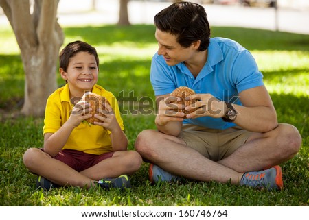 Cute happy boy spending some time with his dad and eating a hamburger in a park