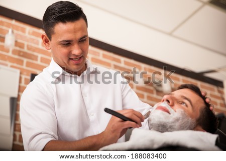 Handsome Latin barber shaving another man\'s beard in a barber shop