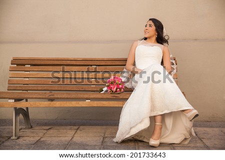 Cute Hispanic bride sitting on a bench outside and looking up towards copy space