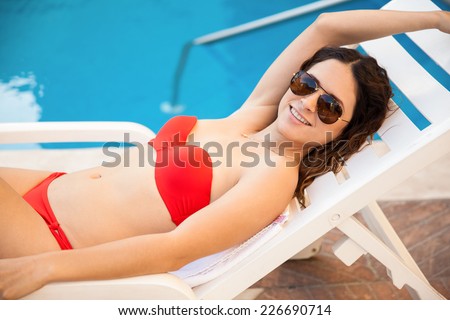 Cute young woman wearing a bikini and sunglasses and relaxing on a poolside chair