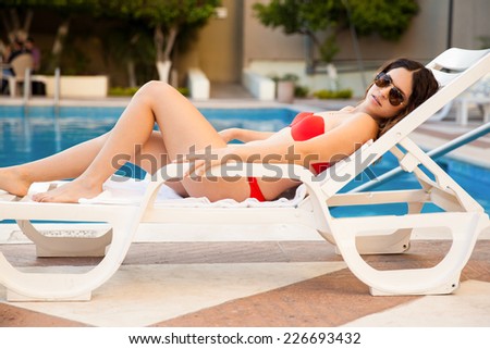 Cute young woman wearing a bikini and sunglasses relaxing on a poolside chair