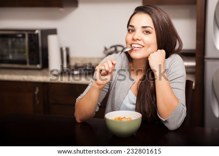 Beautiful girl biting her spoon and smiling while eating some cereal in the kitchen