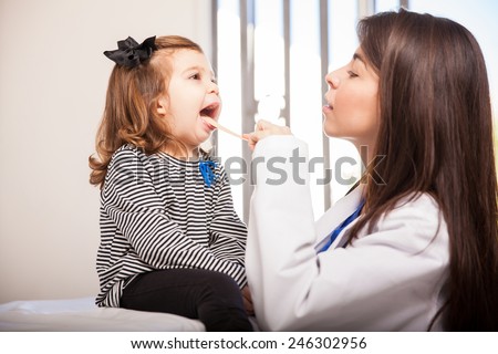 Profile view of a pretty female pediatrician examining a little girl\'s mouth using a stick to hold her tongue down