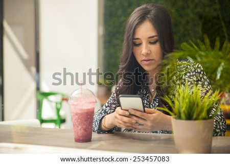 Pretty young brunette using a smartphone while drinking a smoothie in a restaurant