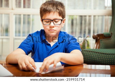 Little boy with glasses folding a sheet of paper into a paper plane at home
